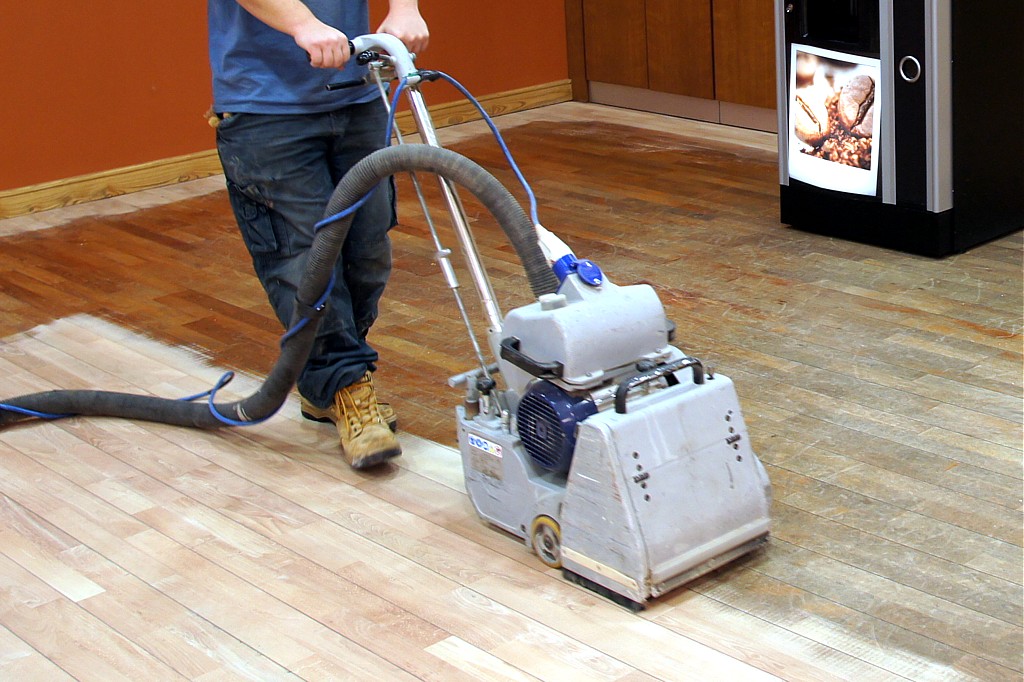 Professional wood floor sanding in progress on a solid maple strip floor, showing a tradesperson operating a drum sander to remove the worn finish.