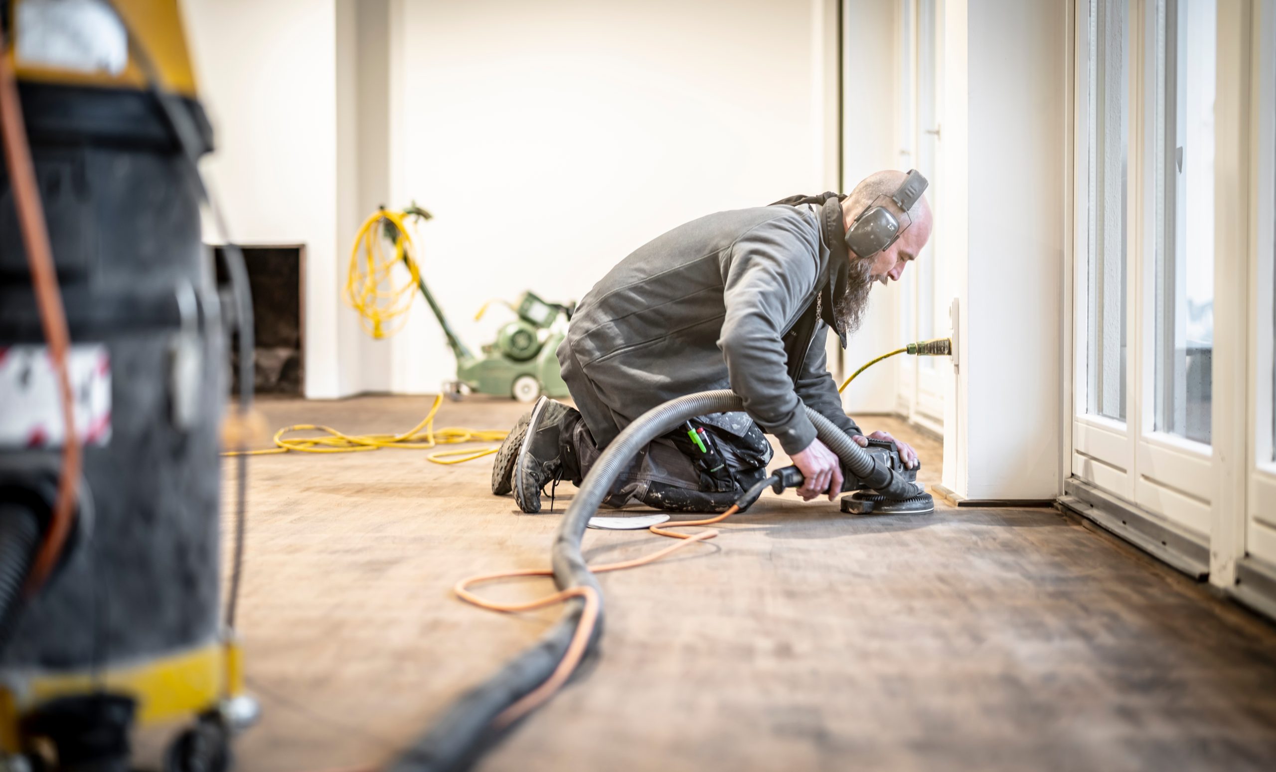Edge sanding during wood floor sanding on a European oak plank floor, showing a professional kneeling with a dust-extraction orbital sander along the skirting.