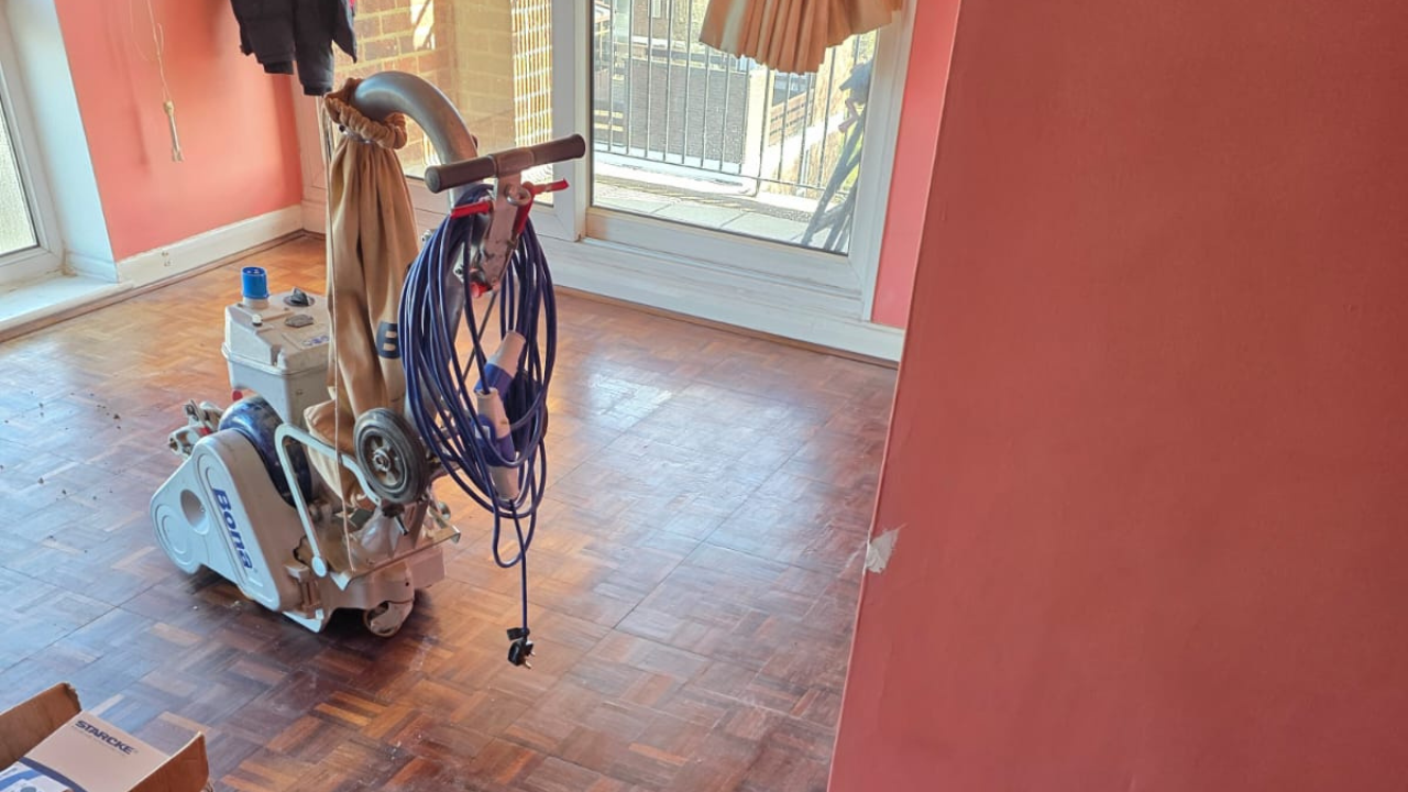 Wood Floor Sanding New Southgate – Oak Parquet Floor in Progress Wood Floor Sanding in New Southgate on an oak parquet block floor, with a professional Bona belt sander in a living room mid-renovation.