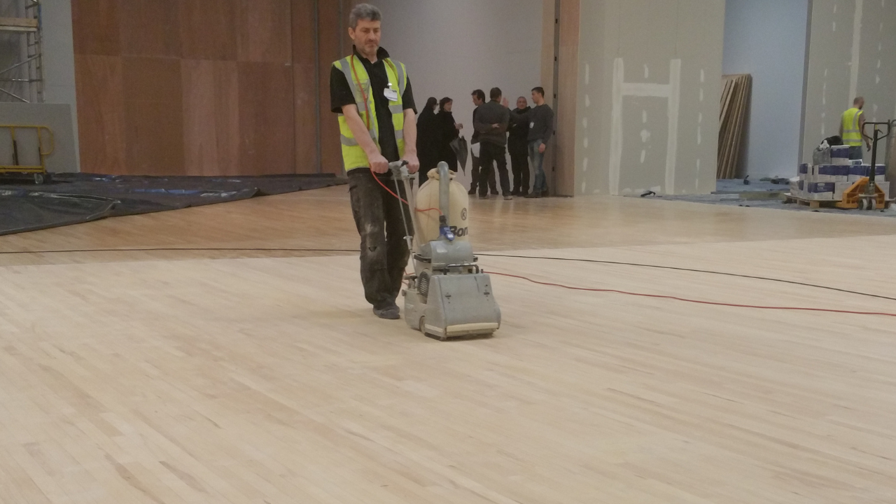 Commercial Floor Sanding New Southgate – Maple Engineered Sports Hall Floor Commercial Floor Sanding in New Southgate on a pale maple engineered sports hall floor, technician using a dust-free belt sanding machine.